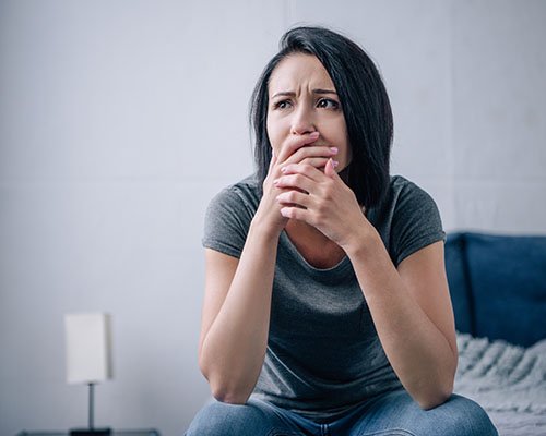 Mujer angustiada, con sus manos cubriendo su boca, sentada al borde de la cama